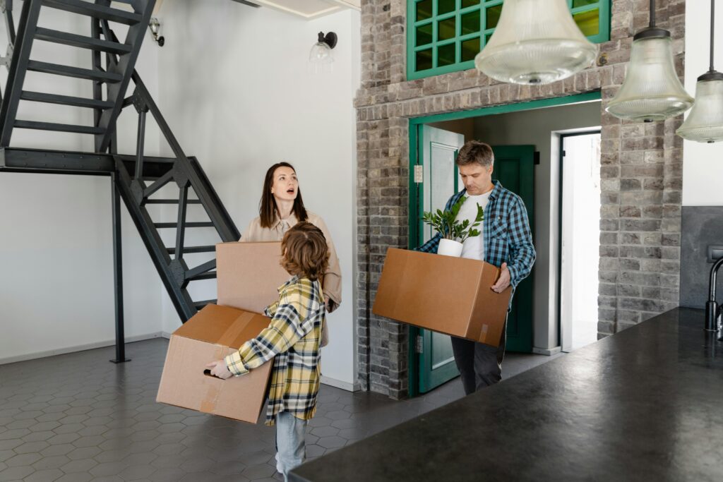 A family moving into their new home, carrying boxes and belongings indoors.